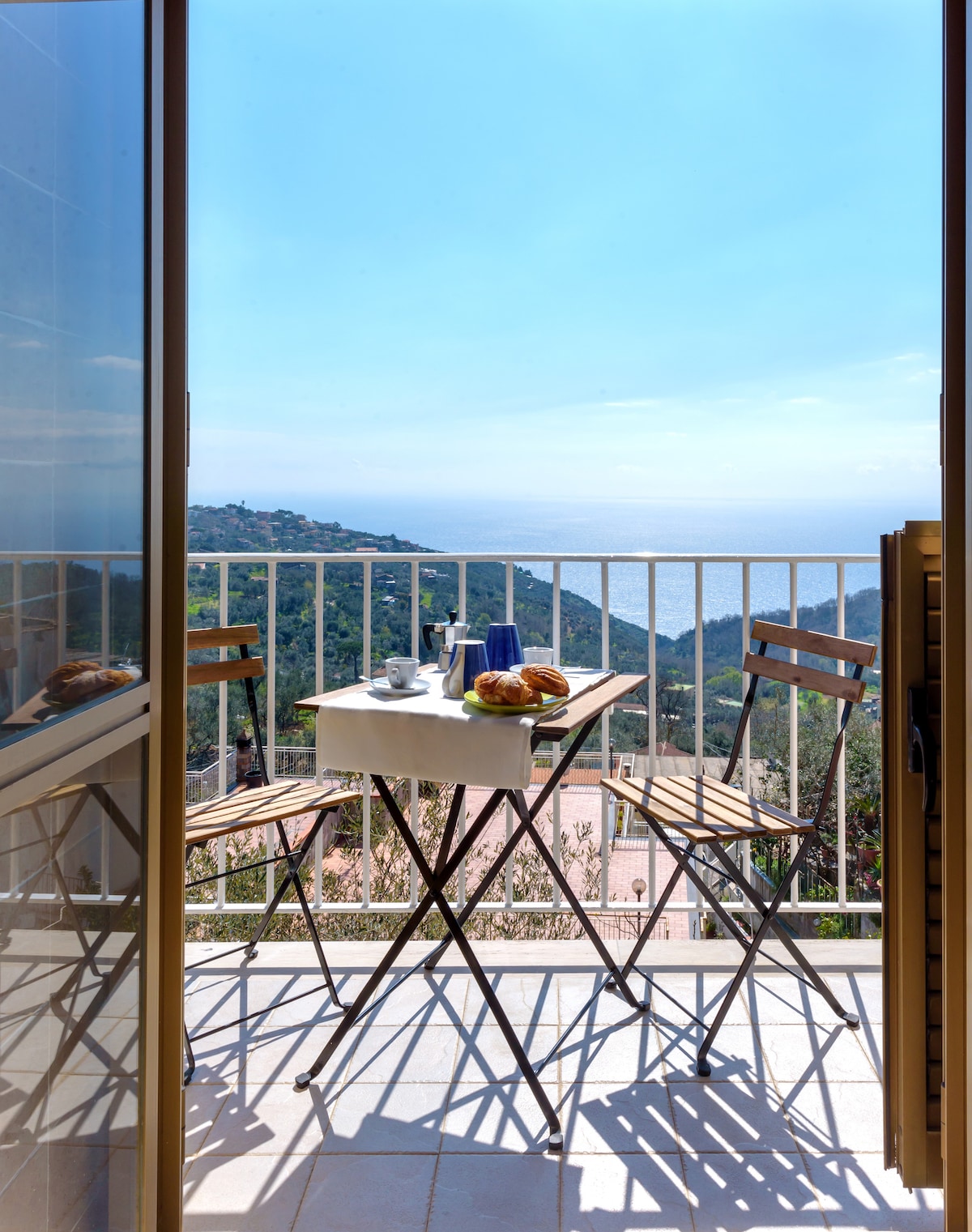 A small balcony is visible, featuring a white table set with beverages and pastries. Two wooden chairs are positioned around the table. The sea can be seen in the background, framed by mountains and a bright sky.