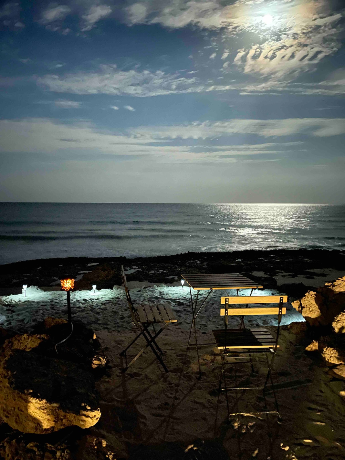A serene beach scene is captured under a luminous moonlit sky. Gentle waves can be seen lapping at the shore, while two folding chairs are positioned on the sandy beach. Soft lighting highlights the surrounding rocks, creating a tranquil ambiance.
