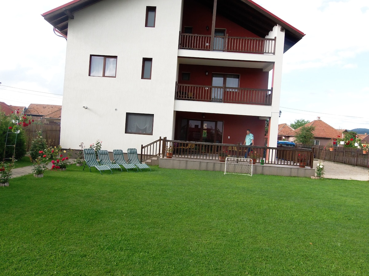 A three-story building is set against a grassy lawn, featuring a balcony on each level. Sun loungers are arranged on the grass, while a small goal is positioned nearby. Flower beds add color to the outdoor space, enhancing the inviting atmosphere.