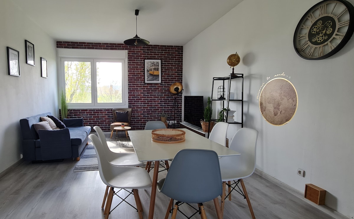 A modern living space features a dining area with a white table and several chairs. A cozy seating arrangement includes a dark blue sofa against a textured brick wall. Natural light enters through a large window, complemented by decorative elements like a clock and greenery.
