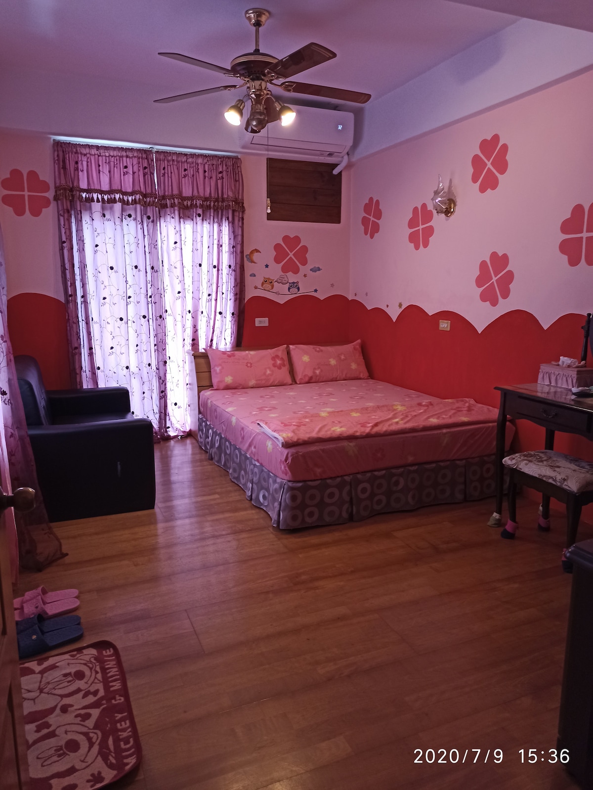 A room features a spacious bed with pink bedding and decorative floral wall accents. Natural light filters through a patterned curtain, complementing the wooden floor. A small seating area and vanity counter are present, creating a functional living space.