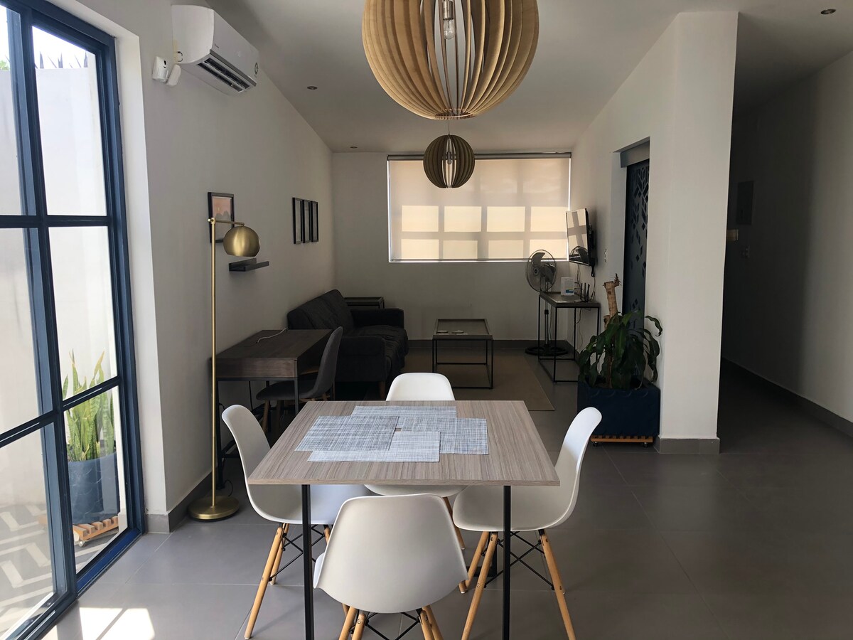A modern dining area features a wooden table surrounded by white chairs. A stylish light fixture hangs above the table. In the background, a cozy seating arrangement with a gray sofa is visible, complemented by a floor lamp and plant accents.