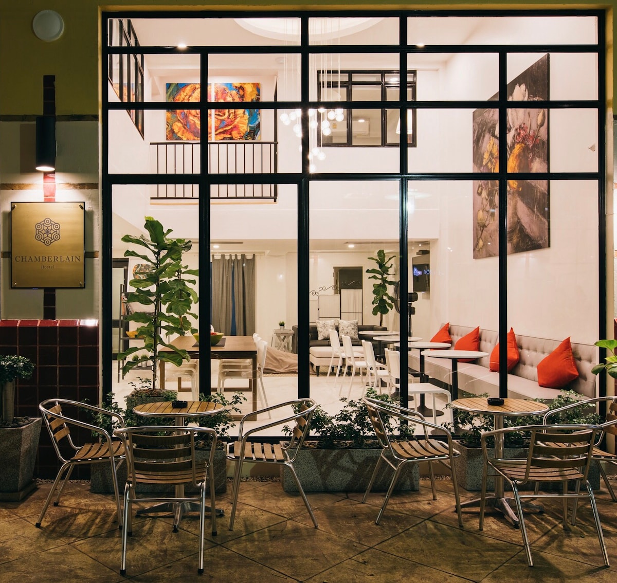The image showcases the exterior of Chamberlain, featuring a large glass façade that allows views of the inviting indoor seating area. Outdoor seating is arranged with metal chairs, surrounded by planters with greenery. Artistic wall decor complements the modern design, creating a welcoming entrance.