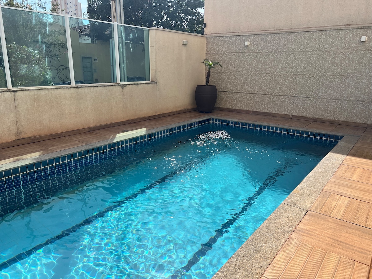 A rectangular swimming pool is framed by natural stone tiles and glass panels, reflecting sunlight across the water's surface. A potted plant is positioned near the pool, adding a touch of greenery to the outdoor space.