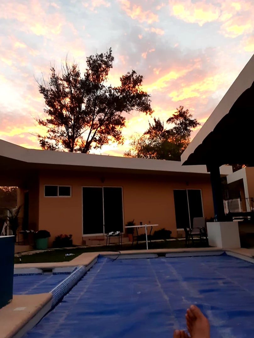 A serene scene captures the sky, adorned with vibrant hues of sunset, framed by trees. The edge of a swimming pool reflects the colorful sky, with the main building visible in the background. Lounge chairs and a table are arranged for relaxation.