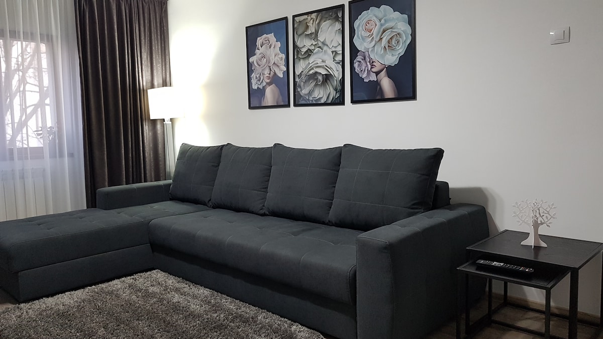 A modern living area features a spacious dark sectional sofa adjacent to a sleek side table. Three floral artworks are displayed above the sofa, complemented by soft lighting from a nearby lamp. Natural light filters through sheer curtains, creating a calm ambiance.
