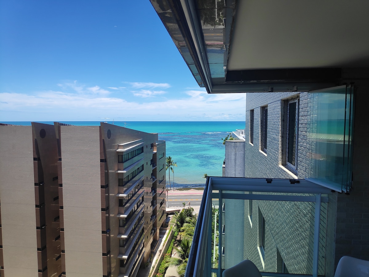 A balcony view shows a clear blue sea meeting the horizon under a bright sky. The landscape includes modern buildings and palm trees, adding a tropical touch. The design of the balcony features glass railings, enhancing the visibility of the surrounding scenery.