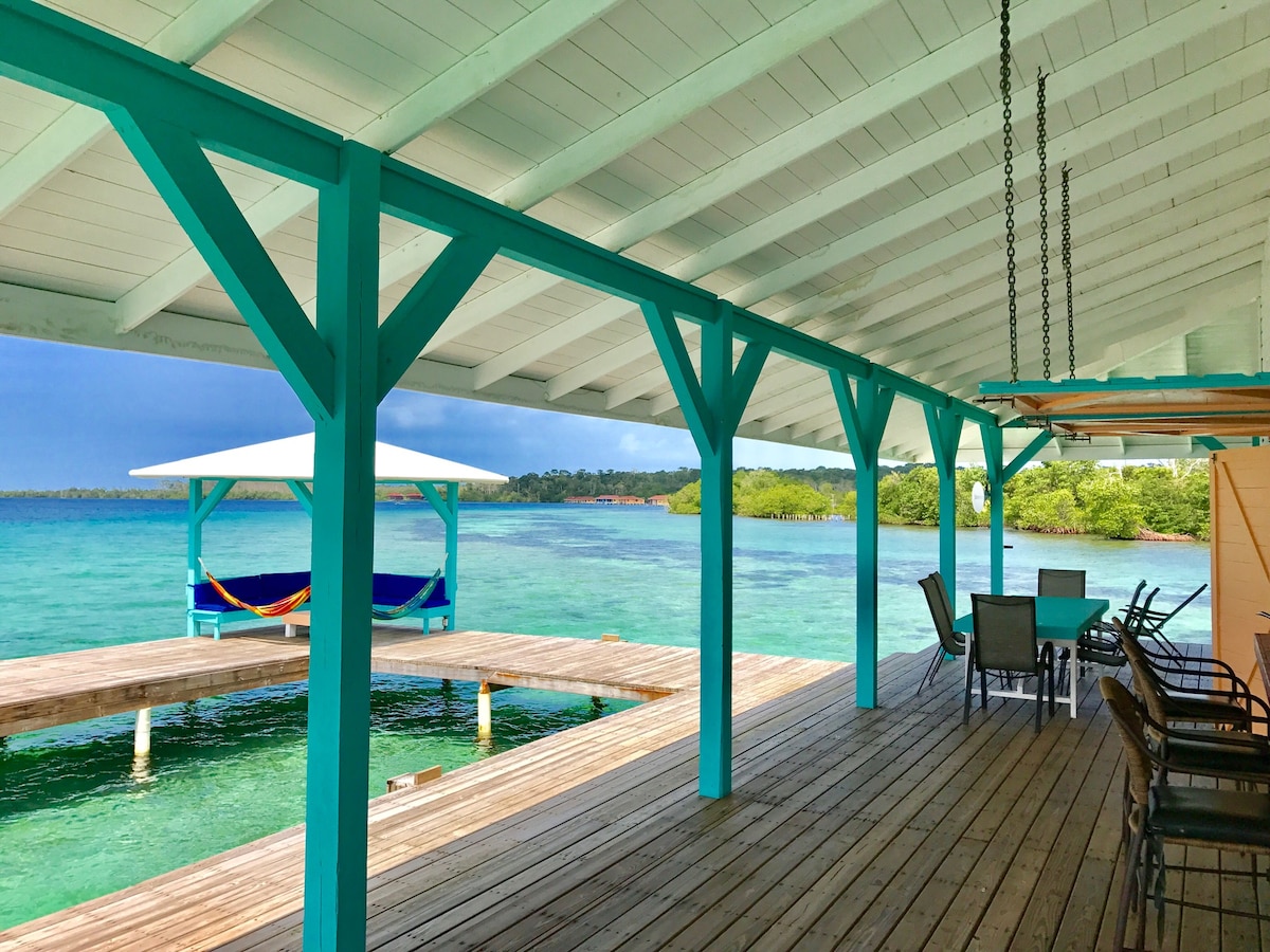 A spacious outdoor deck is supported by teal wooden beams, overlooking calm turquoise waters. A hammock hangs invitingly nearby, while a dining table with several chairs is positioned under the shaded area, creating a relaxed space to enjoy the tranquil surroundings.