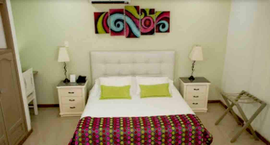 A well-lit bedroom features a spacious bed with white linens and vibrant green accent pillows. Colorful wall art adds a pop of color above the bed. Nightstands with lamps are positioned on either side, and a folding luggage rack is visible in the corner.