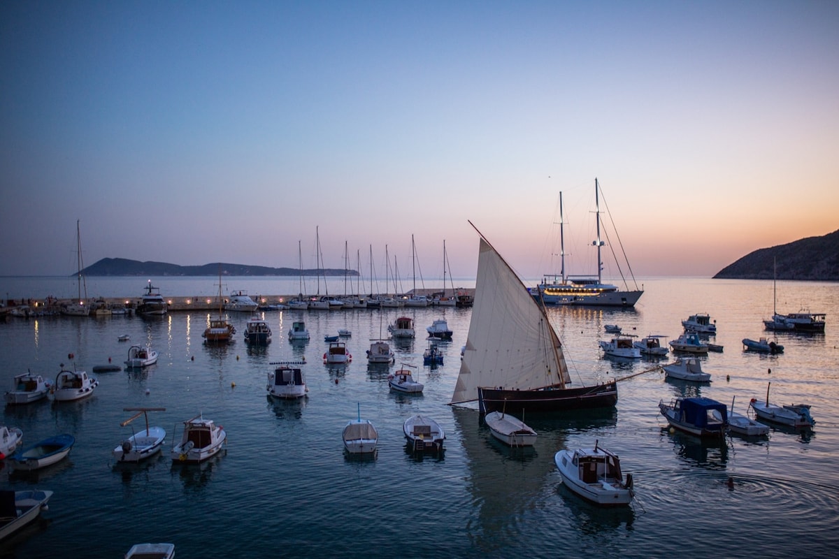 A serene view of a harbor at dusk, featuring a variety of boats bobbing gently in tranquil waters. The horizon displays soft hues of purple and orange, while a sailboat stands prominently with its sail raised. The distant islands add to the peaceful scene.