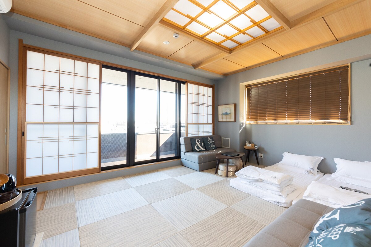 A bright and spacious room features large sliding glass doors with traditional shoji screens, allowing natural light to fill the area. A comfortable sofa is positioned near the window, and several neatly arranged bedding sets are visible on the floor.