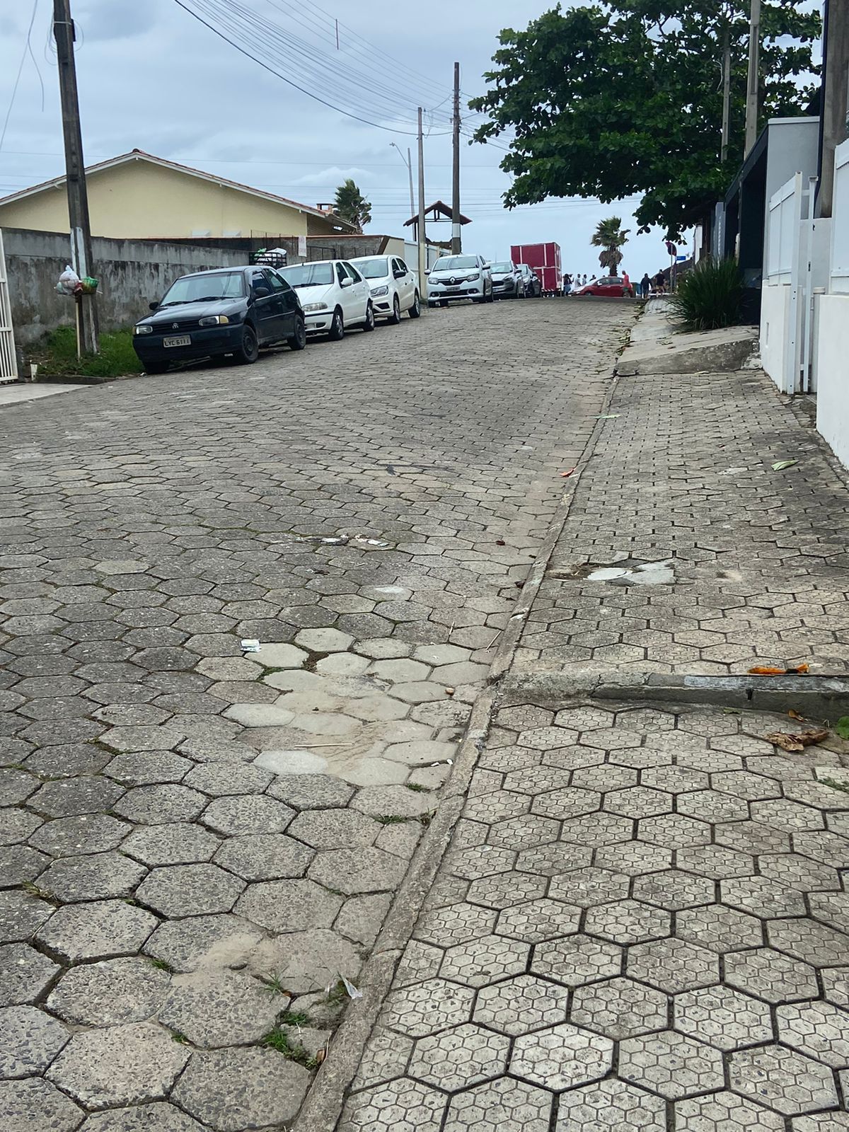 A sloped street is depicted, lined with parked vehicles on both sides. The pavement is made of hexagonal blocks, showing some wear. In the distance, a glimpse of a beach and palm trees can be seen, suggesting proximity to coastal attractions.
