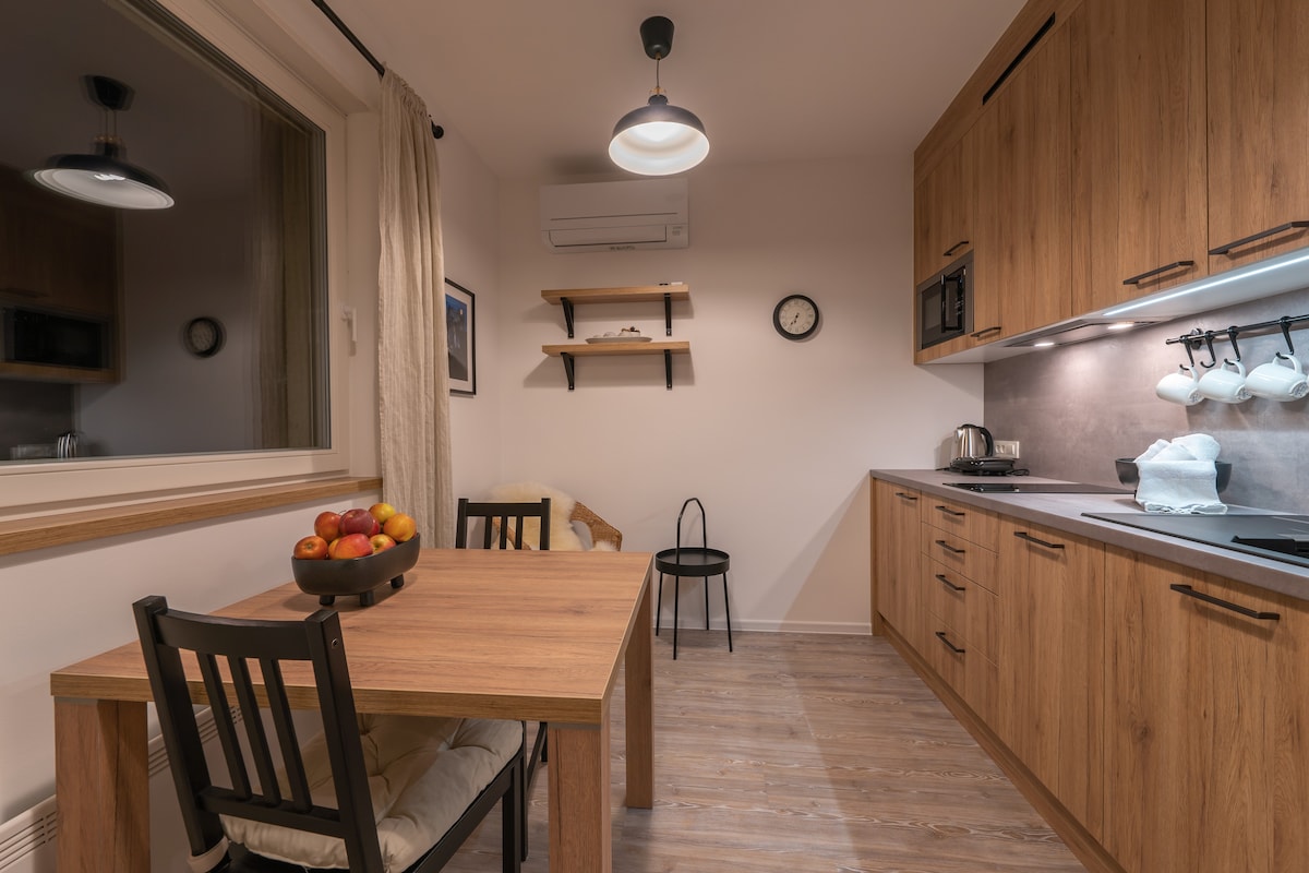 A modern kitchen space features wooden cabinetry and a dining table set for four, accompanied by a bowl of fresh apples. An air conditioning unit and clock are visible on the wall, while a small chair and shelves provide additional functionality.