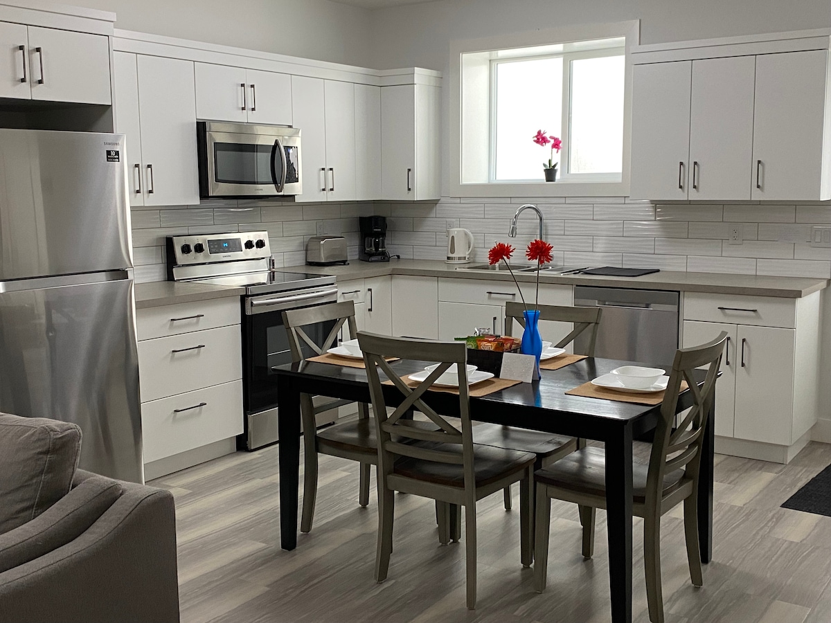 A modern kitchen is equipped with stainless steel appliances, including a refrigerator and oven. A dining table with four chairs is set with place settings. Natural light shines through a window above the sink, where a small vase of flowers adds a touch of colour.