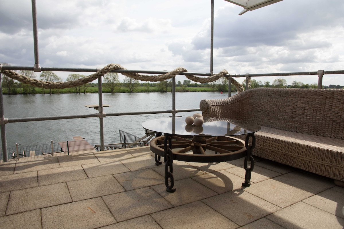 An outdoor terrace features a round glass table with decorative items and a comfortable wicker seating area. The tranquil water view is surrounded by greenery, offering a serene backdrop for relaxation. A railing with nautical rope detail enhances the waterfront experience.
