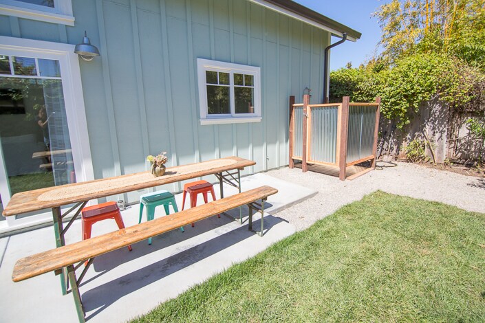 A spacious outdoor area features a wooden picnic table with colorful stools, providing an inviting space for dining. An enclosed outdoor shower stands nearby, surrounded by greenery, enhancing privacy. Fresh grass covers the ground, and the exterior of the house is finished with light blue siding.