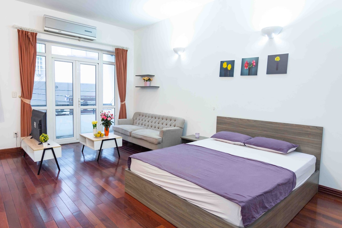 A spacious sleeping area features a large bed with a purple coverlet and light wood accents. A comfortable gray sofa and two tables are positioned nearby. Natural light enters through expansive glass windows draped with sheer curtains, complemented by decorative wall art and fresh flowers.