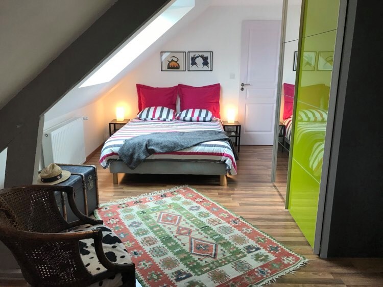 A bright bedroom features a spacious bed with striped bedding against a sloped ceiling. Nightstands with soft lighting flank the bed, while a colorful rug adds warmth to the wooden floor. A woven chair and a decorative hat are placed nearby, enhancing the charm of the space.