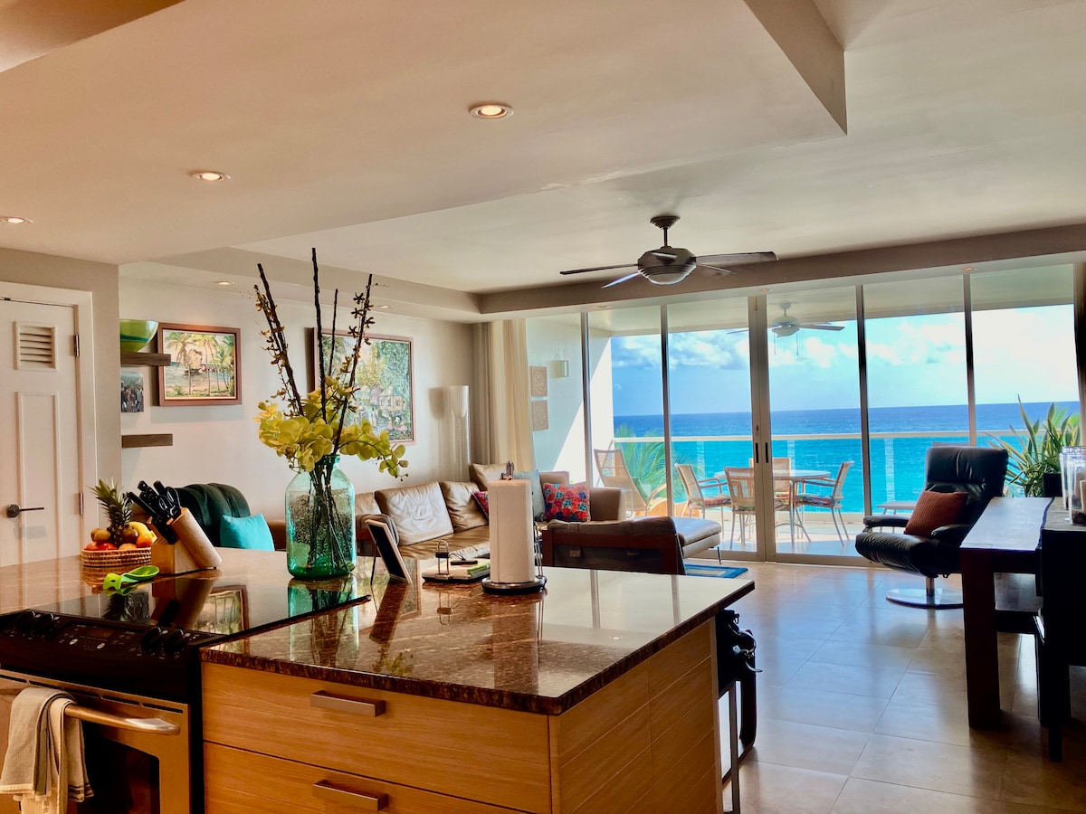 The open-concept living area features a modern kitchen with a large island, showcasing stainless steel appliances and a vase of flowers. Direct views of the coastline are framed by expansive glass doors leading to a spacious balcony with seating.