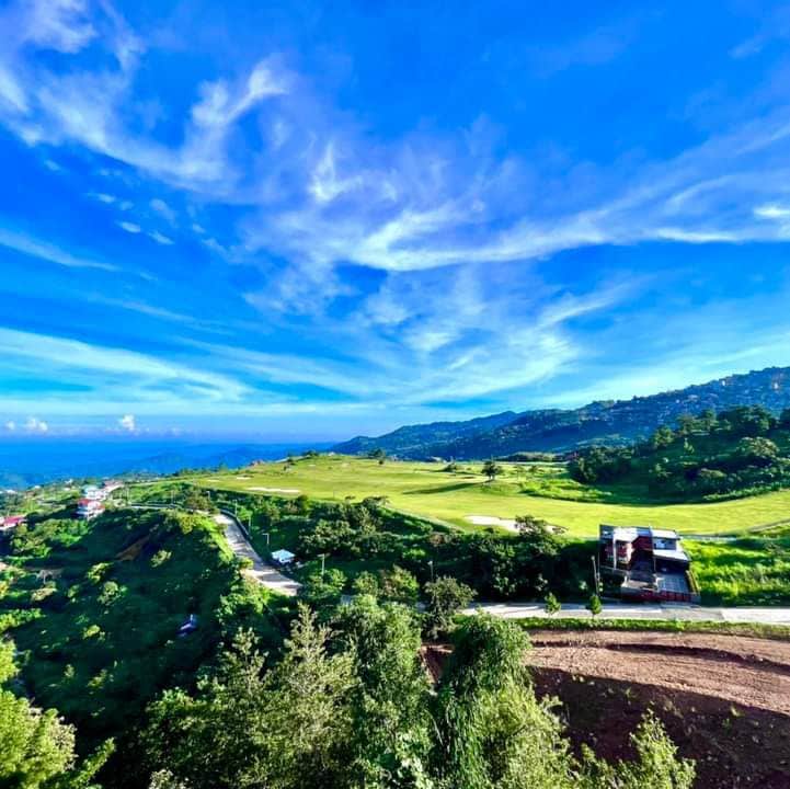 A panoramic view showcases rolling green hills leading towards the ocean. A clear blue sky extends overhead, with wispy clouds enhancing the expansive landscape. The scene captures the natural beauty surrounding the area, indicating a spacious and serene environment.