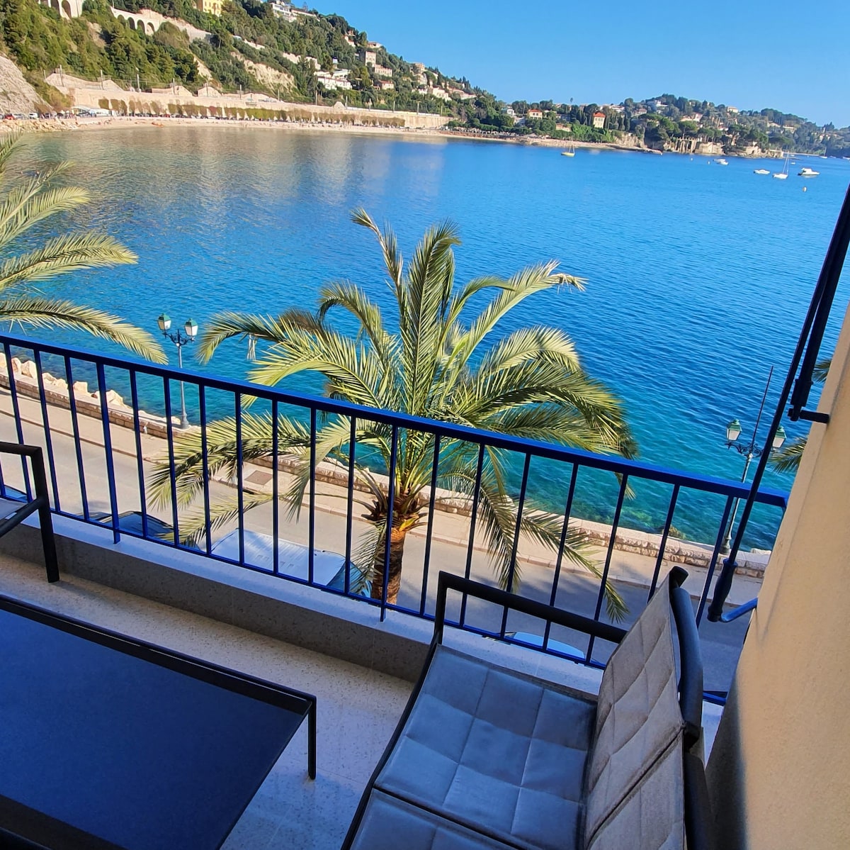 An ocean view is presented from a balcony featuring two chairs and a small table. Palm trees line the waterfront below, while the serene blue sea reflects the sky. The promenade is visible alongside, enhancing the tranquil coastal scenery.