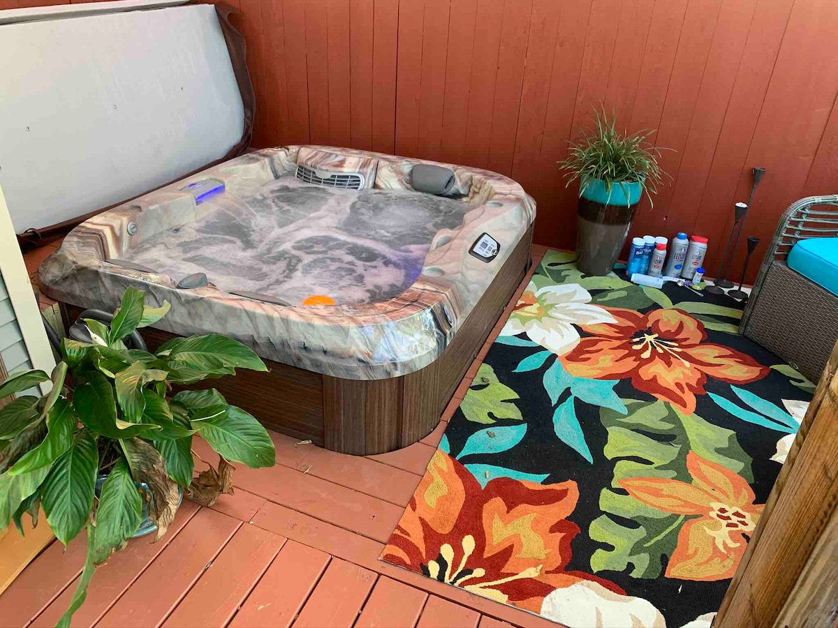 A hot tub with a protective cover sits on a wooden deck, surrounded by colorful floral-patterned outdoor rug and potted plants. Nearby, a small seating area is visible, contributing to a relaxing outdoor space.