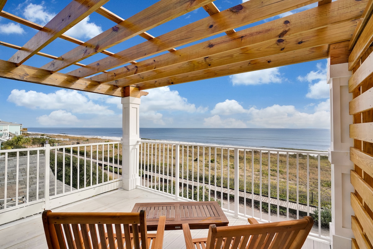 An outdoor deck is showcased with wooden furniture, including a table and chairs, overlooking the vast ocean. A wooden pergola adds architectural interest, complementing the surrounding coastal scenery under a bright blue sky with scattered clouds.