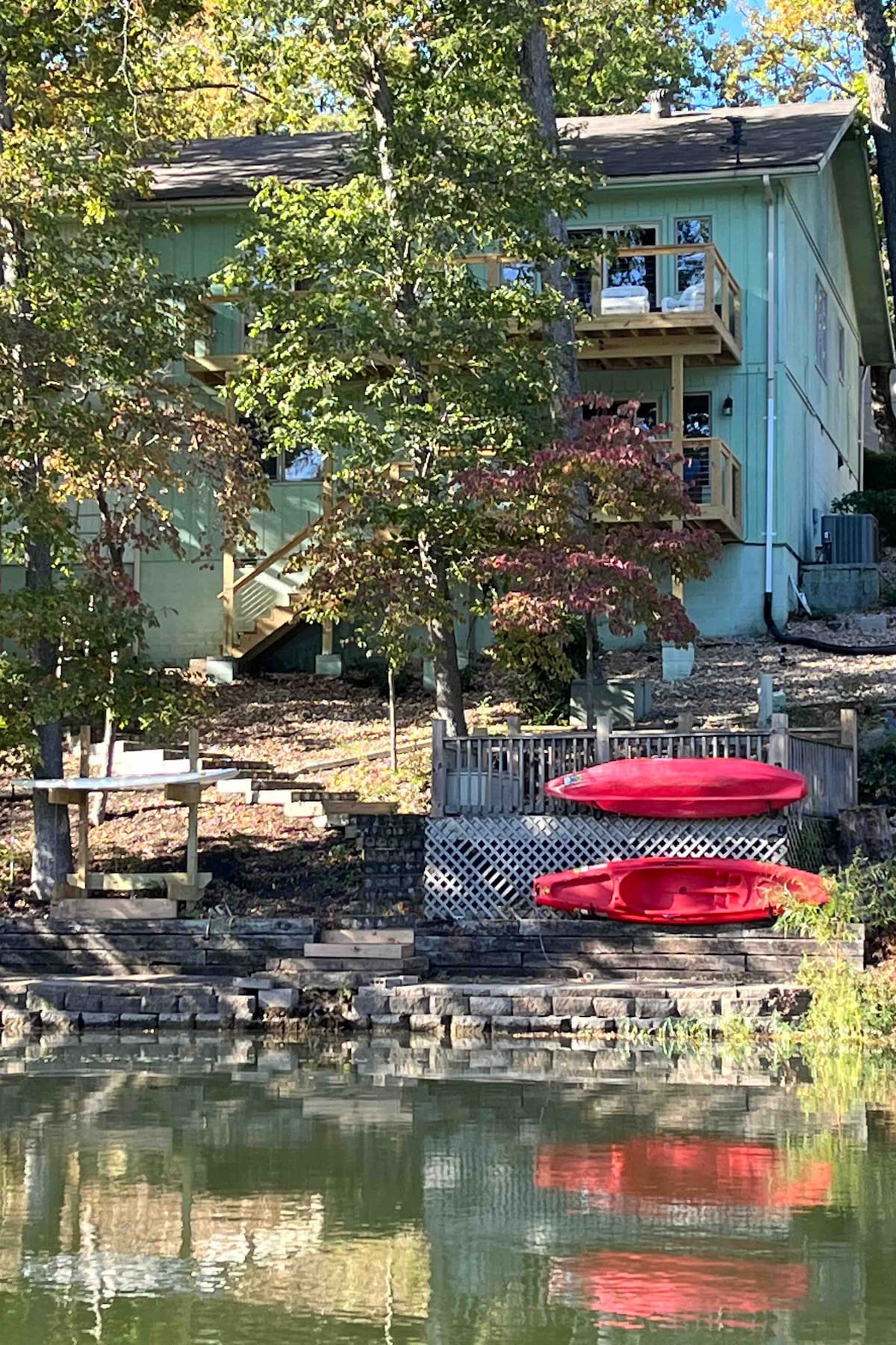 A serene lakeside view features a two-story green townhome surrounded by trees. Two red kayaks are positioned near the water's edge, and a wooden deck is visible, leading to the entrance of the home. The calm lake reflects the colors of the environment.