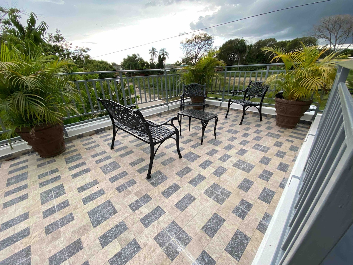 A spacious rooftop terrace is furnished with four metal chairs and two large potted plants. The tiled surface features a checkered pattern in neutral tones, providing an open space for relaxation. Greenery and scenic views are visible beyond the railing, enhancing the outdoor atmosphere.