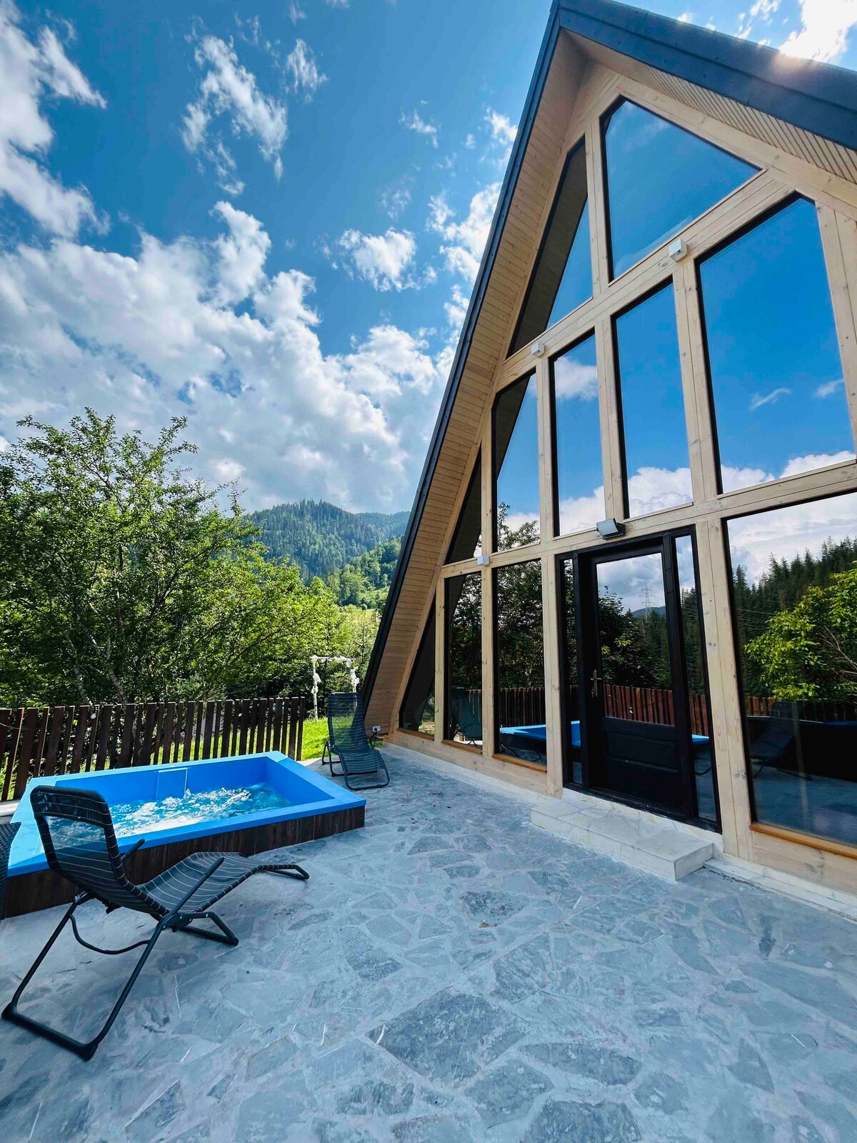 A modern cabin features a striking A-frame design with large glass windows reflecting the surrounding nature. An outdoor jacuzzi is positioned on a stone patio, complemented by black lounge chairs. Lush greenery and scenic mountain views create a serene backdrop.