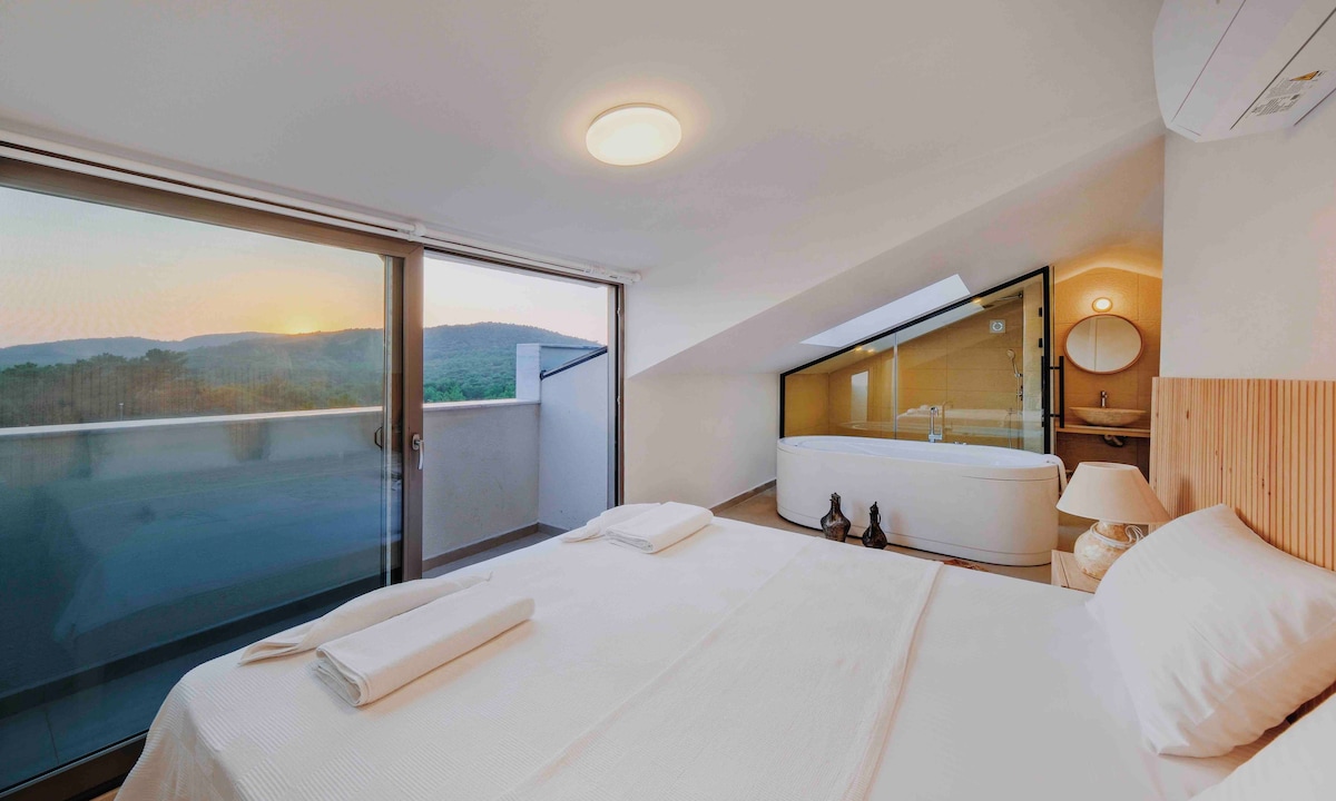 A bright bedroom features a spacious bed with soft linens and towels neatly arranged. Floor-to-ceiling glass doors provide a view of a serene landscape at sunset. A modern bathroom with a freestanding tub and a round mirror is visible in the background.