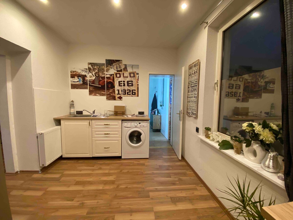 A functional kitchen area features light-colored cabinetry and a washing machine. Wall displays include vintage photographs, adding character. A window allows natural light to fill the space, while a potted plant contributes a touch of greenery to the design.