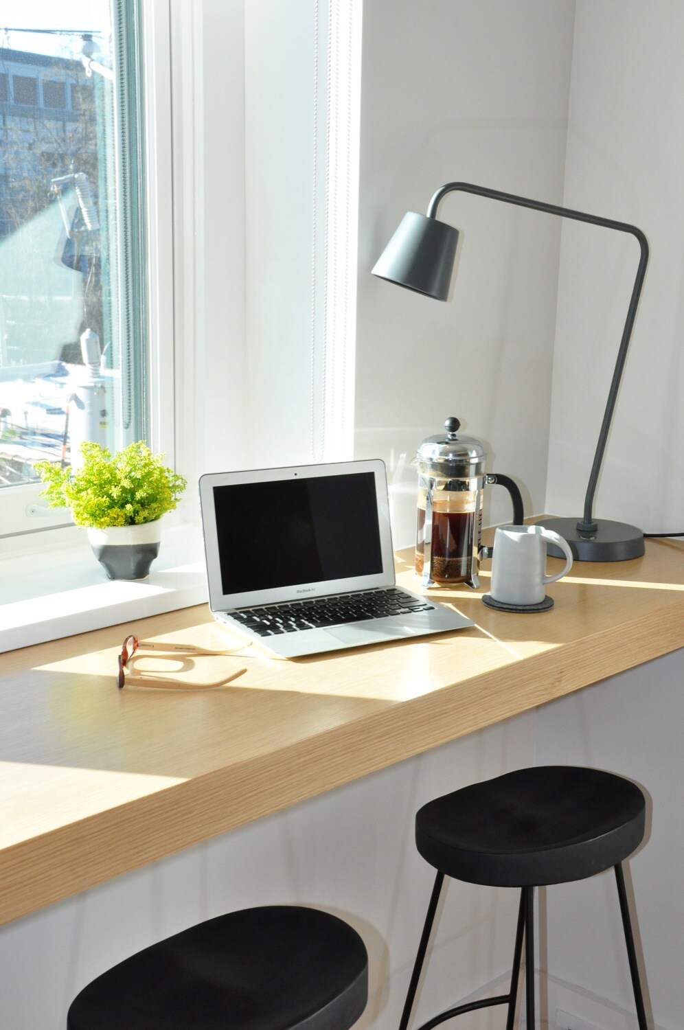 A well-lit workspace is positioned by a window, featuring a laptop on a wooden countertop. A French press and cup accompany the laptop, with a small potted plant adding a touch of greenery. Two black stools are visible, inviting a comfortable seating option.