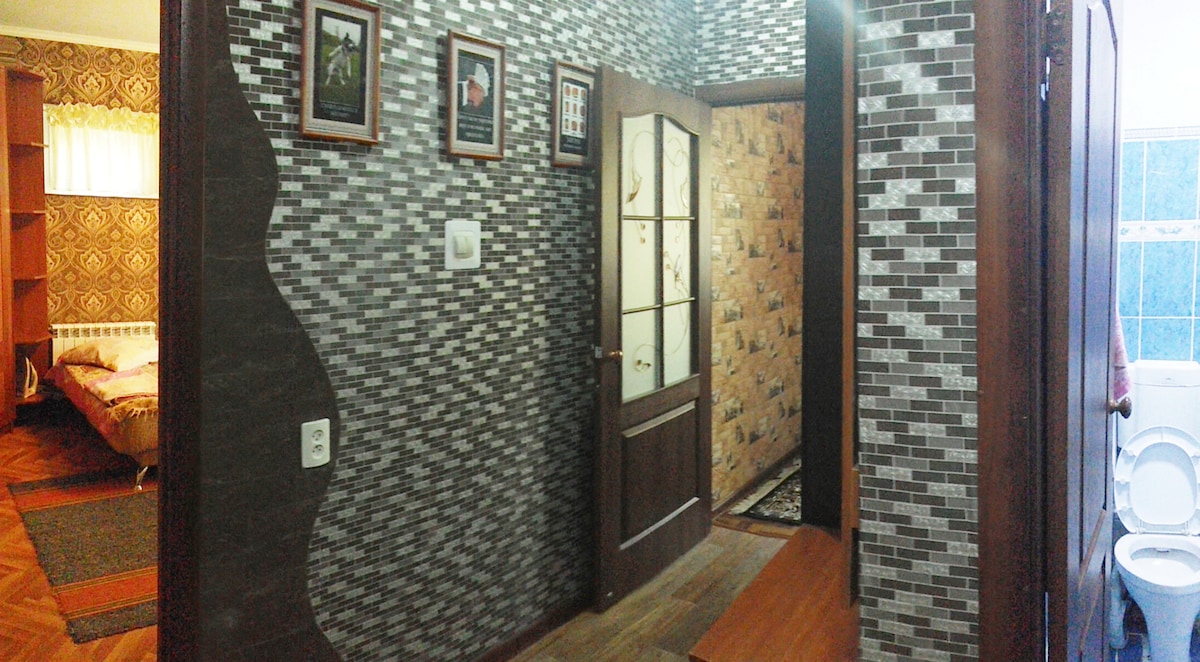 A hallway showcases modern grey patterned walls accompanied by framed photographs. A wooden door leads to another room, while a warm wooden bench is positioned against the wall. Soft natural light filters in from a nearby window, enhancing the welcoming entryway.