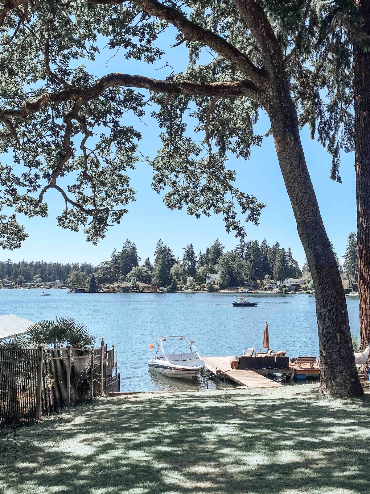 A serene lakeside view is presented, featuring a dock with a boat moored nearby. Tall trees provide shade over a grassy area, while the calm water reflects the surrounding greenery. The distant shoreline is lined with houses, enhancing the peaceful atmosphere.