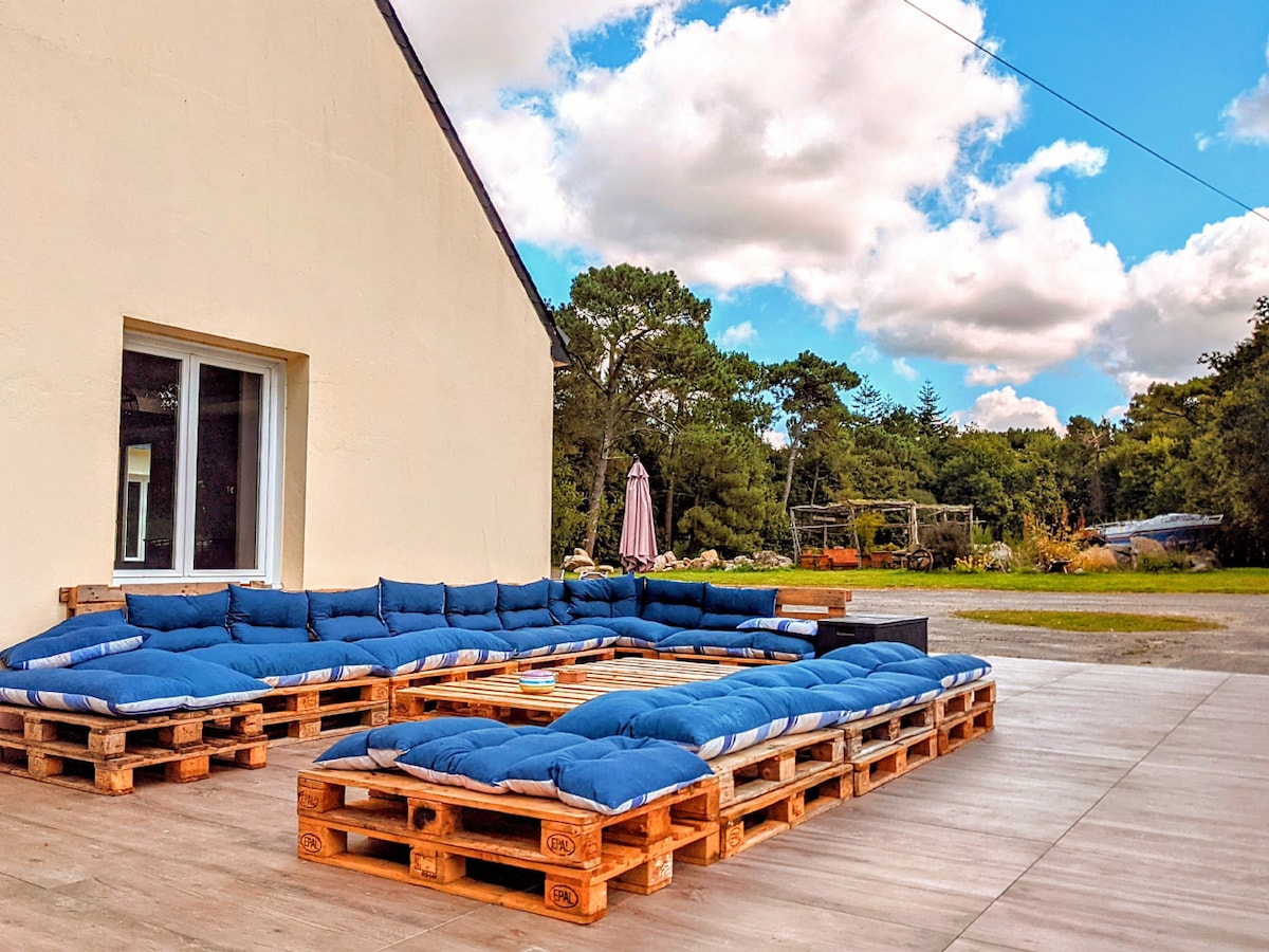 A spacious outdoor seating area is created using wooden pallets, topped with blue cushions for comfort. The setting is surrounded by greenery, with a large umbrella visible nearby. The sky is partly cloudy, enhancing the inviting atmosphere for relaxation.