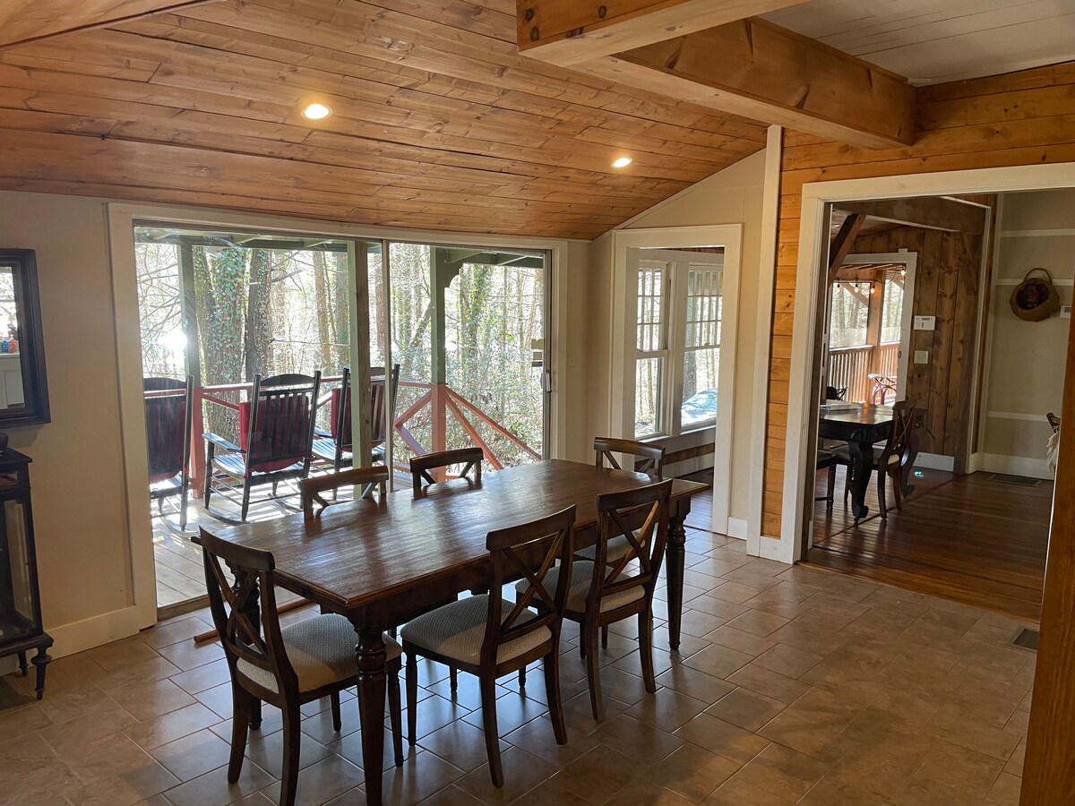 A spacious dining area features a large wooden table surrounded by four upholstered chairs. Natural light filters through expansive sliding glass doors, leading to an outdoor deck. The warm wood ceiling and tiled floor create a welcoming ambiance.