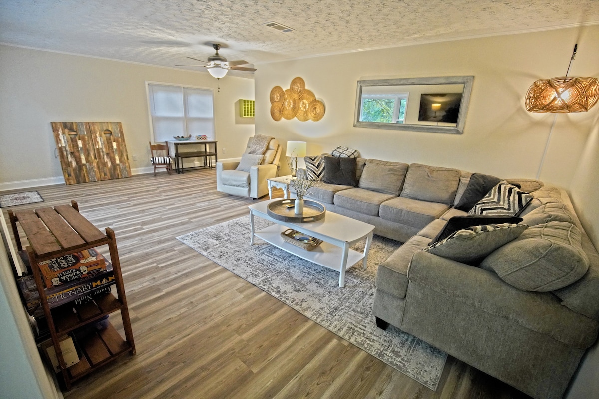 A spacious living area features a large sectional sofa and a coffee table situated on a light rug. A decorative wall art piece hangs beside a window that allows natural light, while a ceiling fan and unique lighting provide a cozy ambiance.