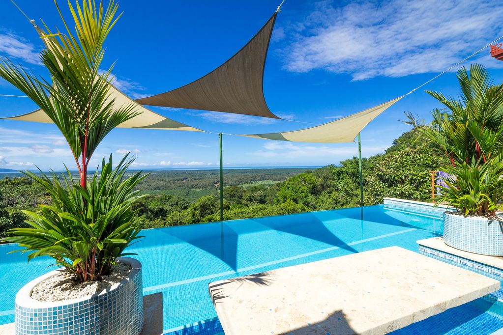 An inviting infinity pool is framed by lush greenery and colorful textile shades. The pool's edge offers breathtaking views of the landscape below, while a submerged stone table provides a unique gathering space for relaxation. Bright blue skies complete the tranquil setting.