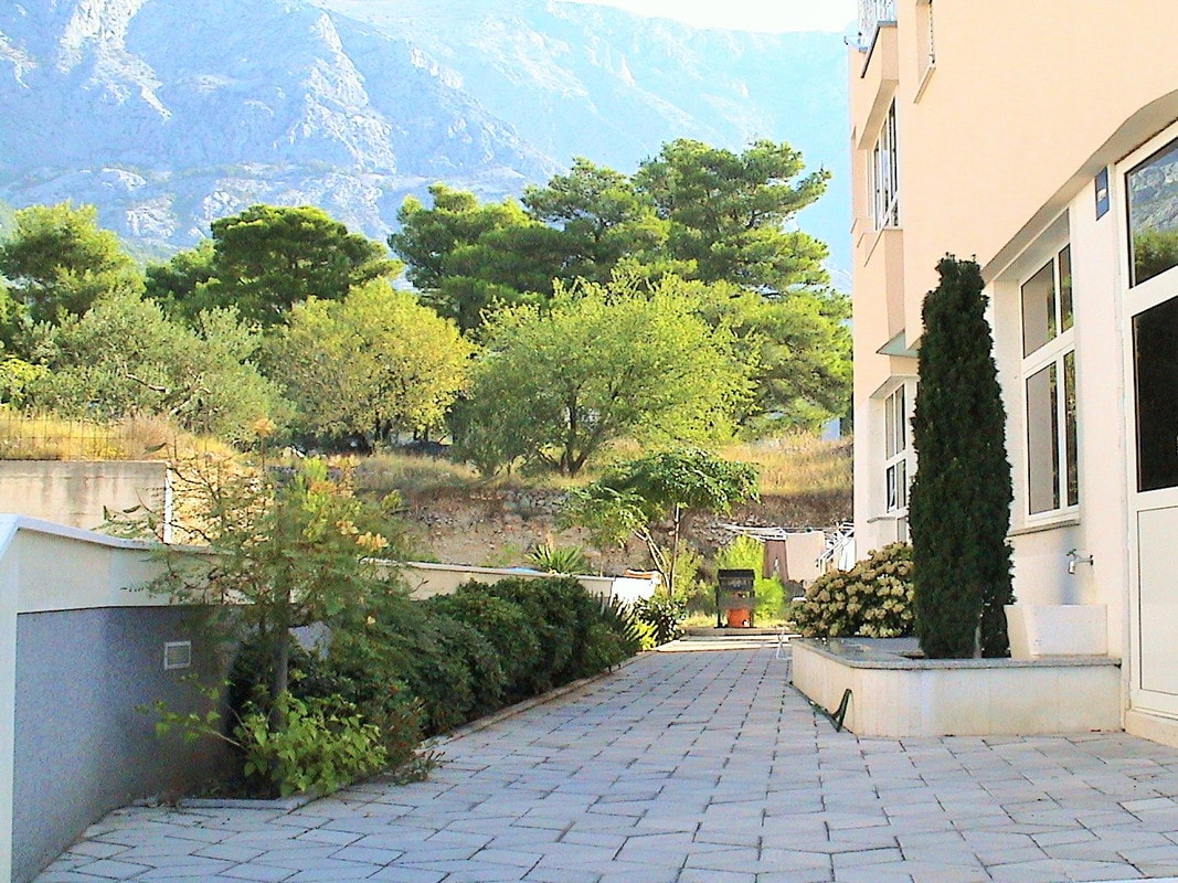 A pathway is lined with greenery, featuring trees and shrubs that blend with the surrounding landscape. The walkway made of stone tiles leads towards a building, with mountains visible in the background. Natural light illuminates the scene, enhancing the tranquil atmosphere.