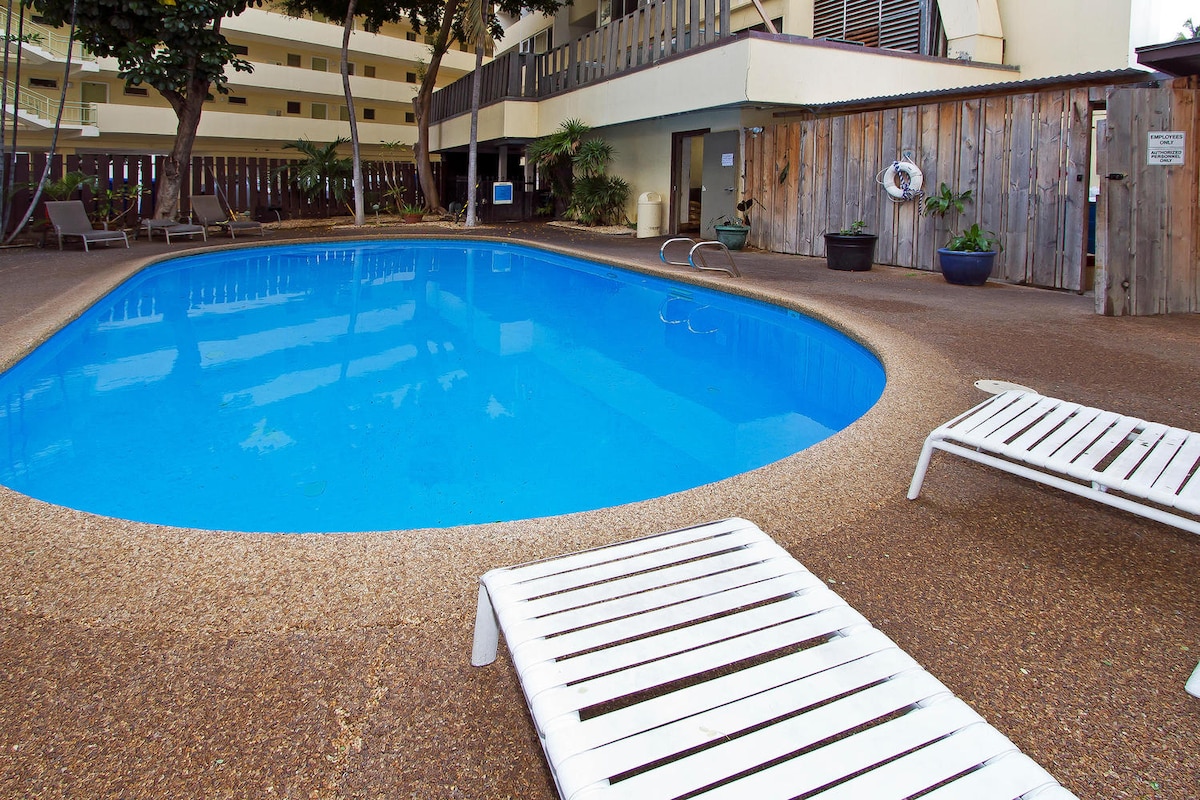 The pool area features a clear blue swimming pool surrounded by a textured concrete deck. Two white lounge chairs are placed nearby, inviting relaxation. Lush greenery and wooden fencing provide a sense of privacy and tranquility.