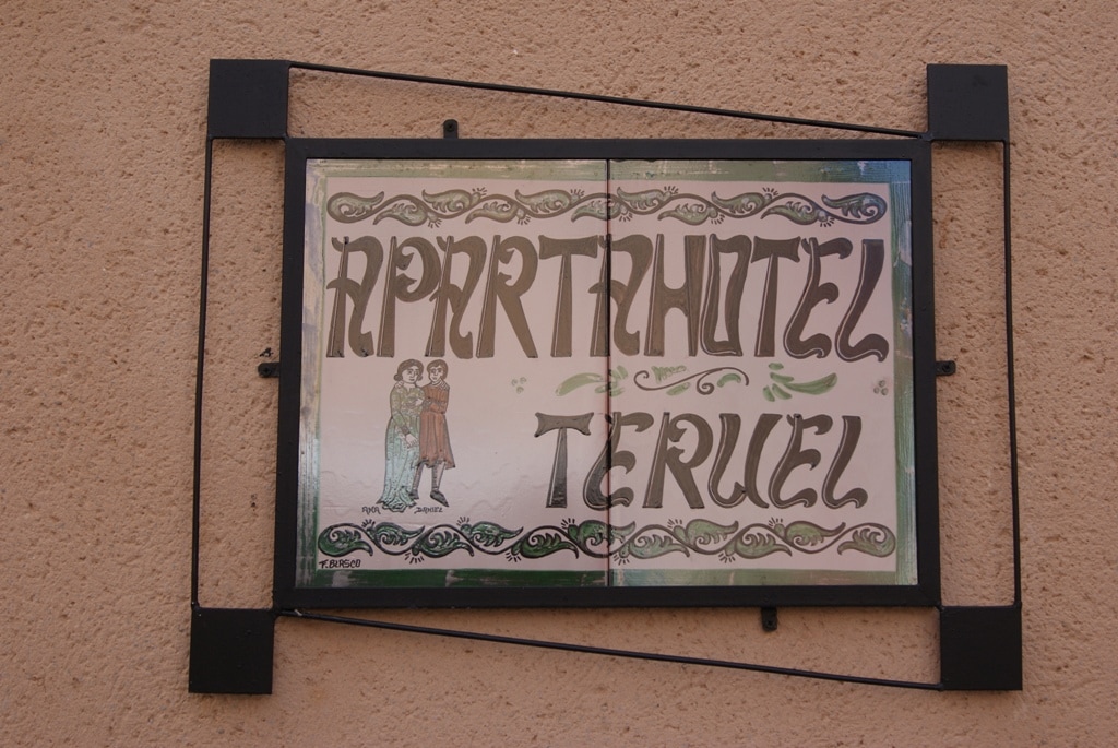 A decorative sign displaying the name 'Apartahotel Teruel' is mounted on a textured wall. The sign features a traditional design with intricate detailing and two figures in the artwork. The colors are softly blended, contributing to the overall aesthetic of the establishment.