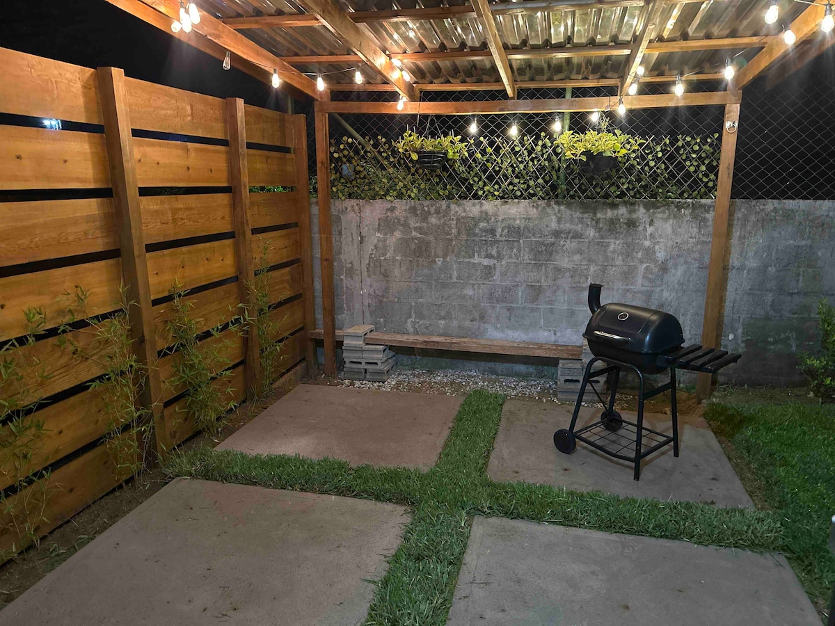 An outdoor space features a wooden pergola adorned with string lights, providing a cozy setting. A charcoal grill is positioned on a paved patio area, surrounded by grass and a wooden fence, ensuring privacy. The environment offers potential for relaxation and gatherings.