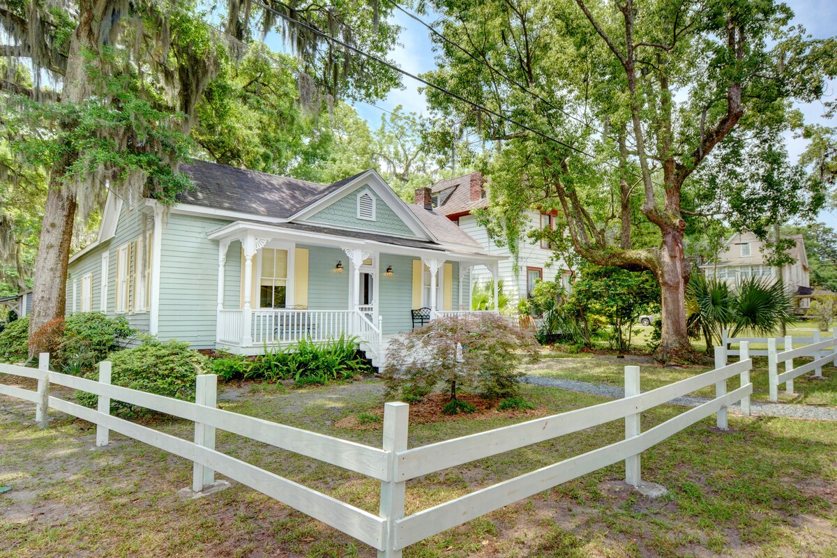 A charming bungalow is surrounded by lush greenery and tall trees. The exterior features light-colored siding with decorative white trim. A covered front porch is highlighted by white railings, and a small garden area is visible in the fenced yard.