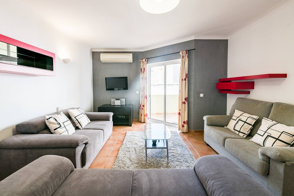 A spacious living area is furnished with two comfortable sofas and a glass coffee table. A wall-mounted television is visible alongside a stylish shelf. Natural light enters through large sliding doors, which lead to a balcony. Neutral tones and light patterns enhance the inviting ambiance.