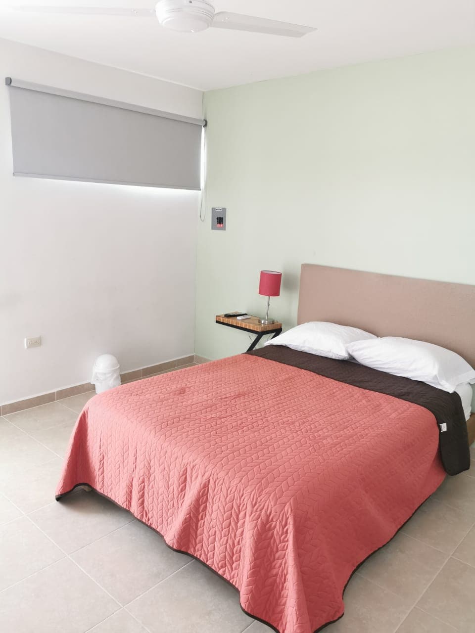 A bright and simple bedroom features a spacious bed with a coral-colored blanket and white linens. A small bedside table holds a lamp, and a fan is mounted on the ceiling. Natural light filters through a window covered with a gray roller shade.