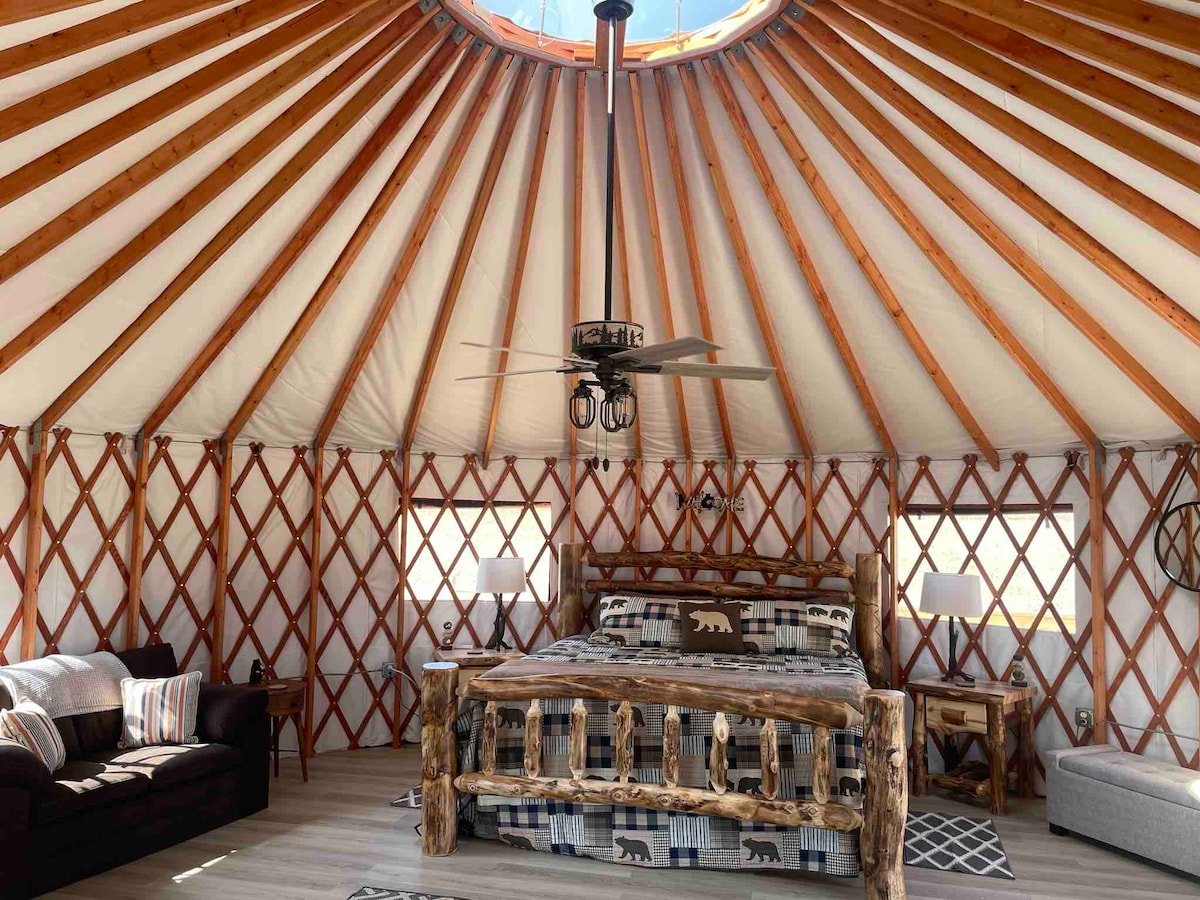 The interior of a yurt showcases a circular design with wooden beams and a central ceiling opening. A large bed made of natural wood features a bear-themed blanket. A cozy seating area includes a dark sofa and a light-colored chair, with light streaming in through two windows.