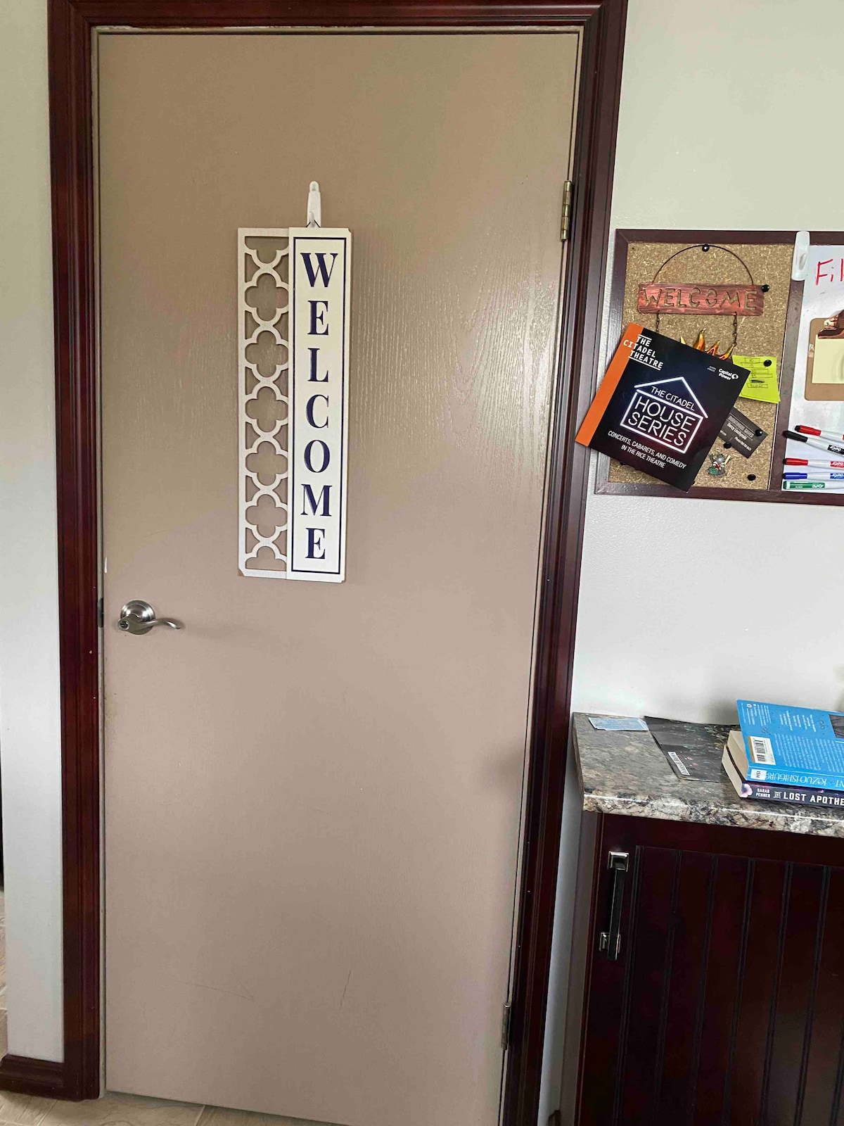 A beige door is displayed with a decorative 'WELCOME' sign hanging from the top. Nearby, a cork bulletin board features various papers and notes, along with a small cabinet showcasing a stone countertop.