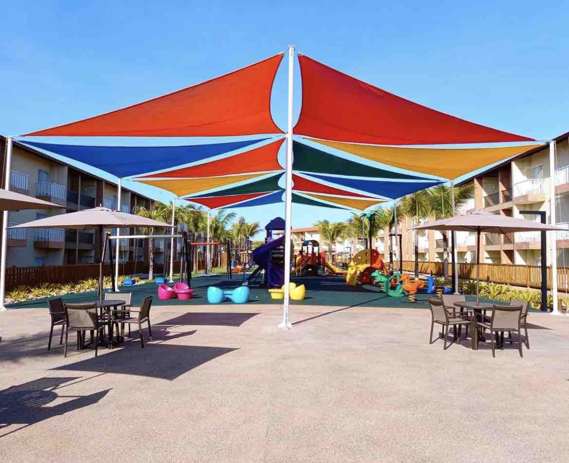 A vibrant play area is shaded by colorful canopies, featuring a playground with slides and climbing structures. Outdoor seating is provided with several tables and chairs, creating a welcoming space for relaxation. Lush greenery lines the perimeter, enhancing the outdoor environment.