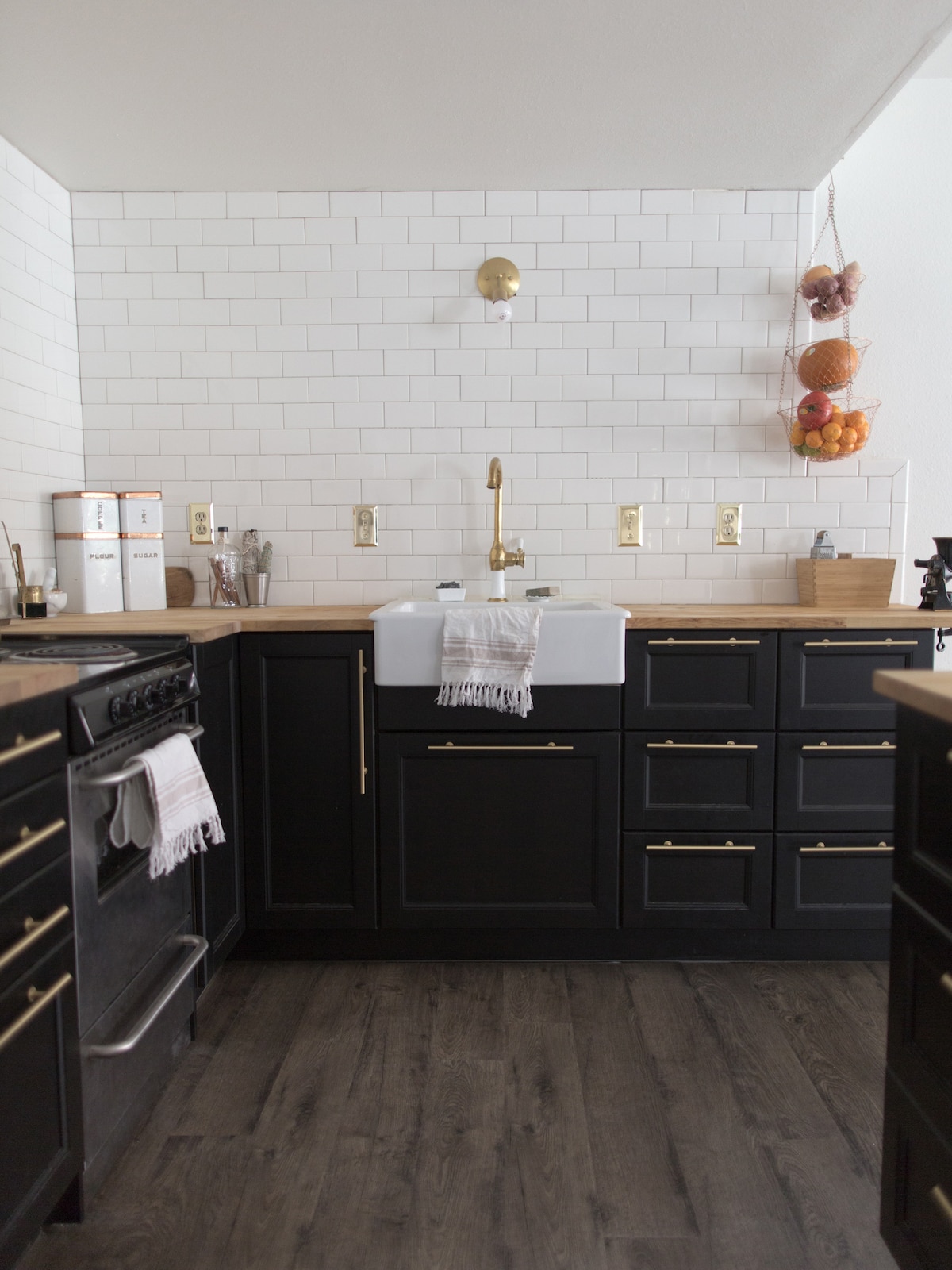 A modern kitchen features black cabinetry with gold hardware and a spacious farmhouse sink. Subway tiles create a clean backdrop, while wooden countertops provide warmth. A stovetop and wall-mounted shelving are included, showcasing kitchen essentials and decorative items.